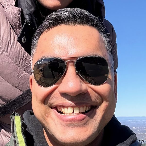 Sid Harsha smiling outdoors on a mountain hike, wearing sunglasses